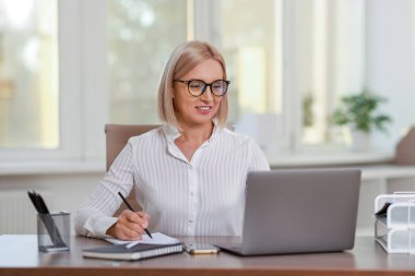 Ofiste dizüstü bilgisayarla çalışan gülümseyen orta yaşlı kadın.