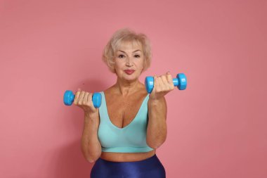 Üst düzey kadın pembe arka planda dambıllarla egzersiz yapıyor.