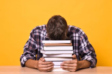 Kitap yığını olan genç bir öğrenci sınavdan önce sarı arkaplan karşısında stres altında.