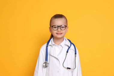 Steteskoplu küçük kız turuncu arka planda doktor gibi davranıyor. Geleceğin mesleğini hayal etmek