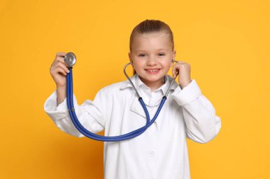 Steteskoplu küçük kız turuncu arka planda doktor gibi davranıyor. Geleceğin mesleğini hayal etmek