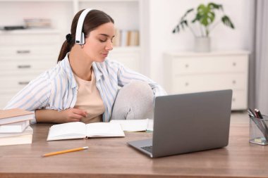 Kulaklıklı öğrenci içeride ahşap masada ders çalışıyor.