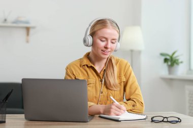 Interpreter in headphones taking notes while working with laptop at table indoors clipart