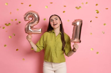 Reşit olma partisi - 21. yaş günü. Pembe arka planda balonları ve konfetisi olan mutlu genç bir kadın.