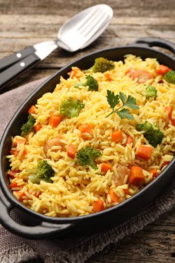 Pişirme kabında sebzeli pilav ve ahşap masada çatal bıçak, yakın plan.