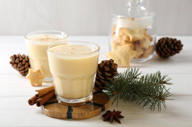 Lezzetli Noel kokteyli (eggnog) bardaklarda, kozalaklarda ve beyaz ahşap masadaki baharatlarda, yakın plan