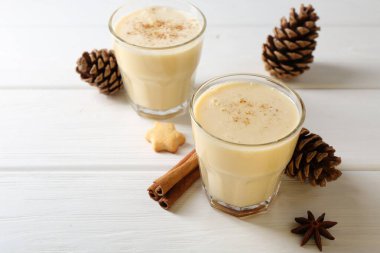 Lezzetli Noel kokteyli (eggnog) bardaklarda, kozalaklarda ve beyaz ahşap masadaki baharatlarda, yakın plan. Metin için boşluk