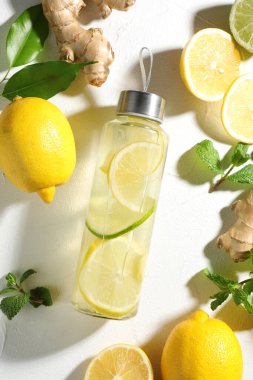 Şişe içinde naneli limon suyu, taze yapraklar, beyaz masada meyve ve zencefil.