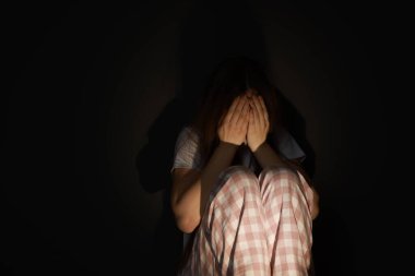 Fear of darkness. Scared young woman sitting on dark background clipart