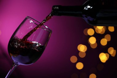 Pouring red wine into glass against purple background, closeup. Bokeh effect clipart