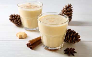 Lezzetli Noel kokteyli (eggnog) bardaklarda, kozalaklarda ve beyaz ahşap masadaki baharatlarda, yakın plan