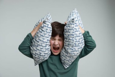 Annoyed boy covering his ears with pillows due to loud sound on grey background clipart