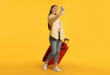 Bavullu mutlu kadın turuncu arka planda selfie çekiyor.