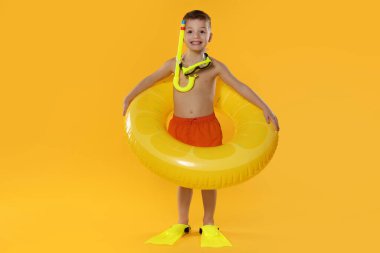 Cute little boy in beachwear with snorkeling equipment and inflatable ring on orange background