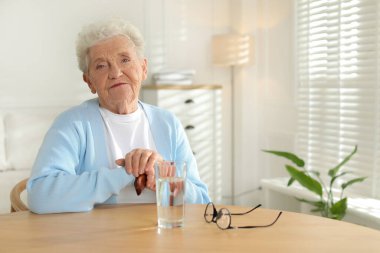 Düşünceli, son sınıf öğrencisi kadın kendini evde bir bardak suyla masada yalnız hissediyor.