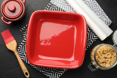 Red ceramic casseroles, baking parchment paper, raw pasta and brush on black table, flat lay clipart