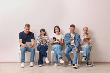 Beyaz duvarın yanındaki sandalyelerde farklı aletler kullanan bir grup insan. Modern teknoloji