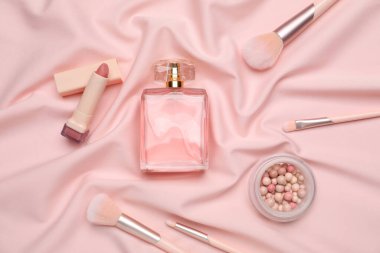 Pembe ipek kumaş üzerinde bir şişe parfüm, top pudrası, ruj ve makyaj fırçası.