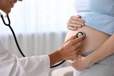 Hamilelik kontrolü. Steteskoplu gülümseyen doktor hastanın karnındaki kalp atışlarını dinliyor.