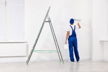 Erkek tamirci duvarları boyuyor, merdiveni kapalı, arka manzaralı.
