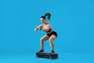 Spor elbiseli kadın egzersiz yapıyor. Açık mavi arka planda adım platformu var. Metin için yer var.