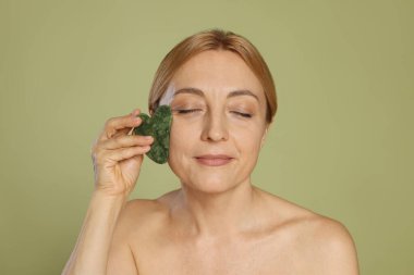 Yeşil arka planda gua sha aracıyla yüz masajı yapan güzel bir kadın.