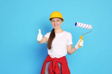 Açık mavi arka planda baş parmaklarını gösteren boya silindirli genç dekoratörün portresi