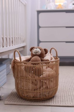 Çocuk odasındaki beşiğin yanında battaniye ve oyuncak ayıyla dolu bir sepet.