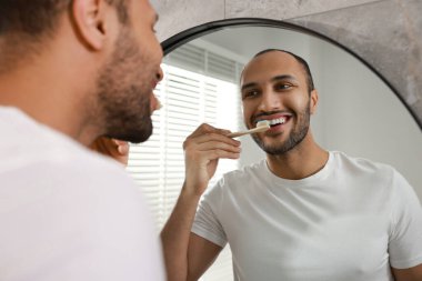 Gülümseyen adam banyoda aynanın yanında dişlerini fırçalıyor.