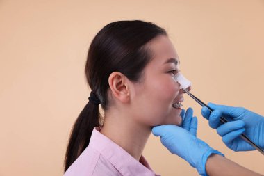 Doctor removing medical bandage from patient's nose after plastic surgery operation on beige background, closeup clipart