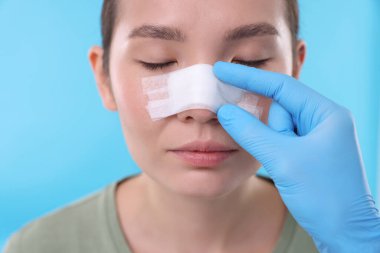 Doctor checking patient's nose after plastic surgery operation on light blue background, closeup clipart