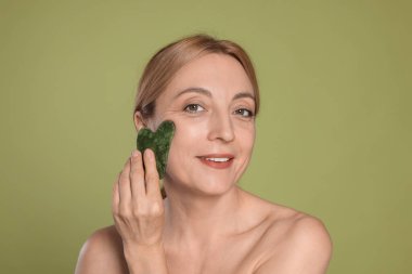 Gülümseyen kadın yeşil arka planda gua sha aracıyla yüz masajı yapıyor.