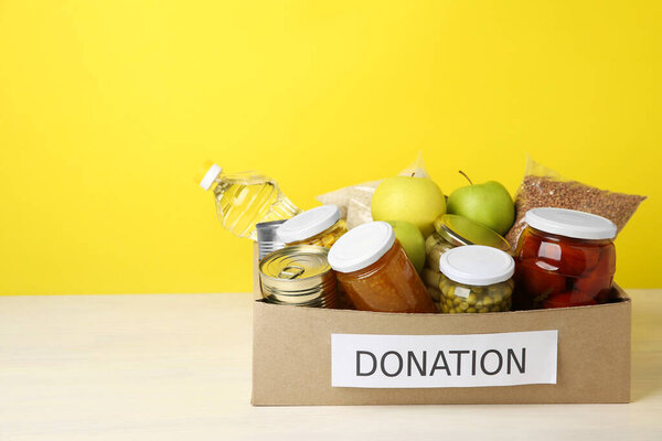 Различные пищевые продукты для пожертвования в коробке на столе на желтом фоне, пространство для текста