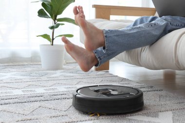 Genç adam dizüstü bilgisayar kullanırken elektrikli süpürge evde halı süpürüyor.