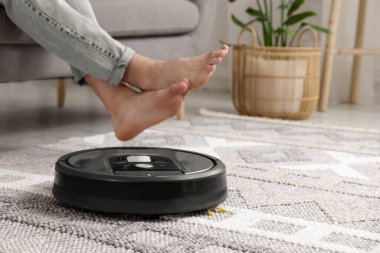 Genç adam kanepede dinlenirken robotik elektrikli süpürge evdeki yerleri süpürüyor.