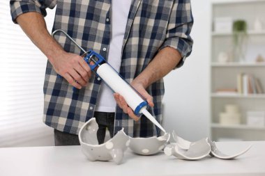 Man with caulking gun glueing pieces of ceramic piggy bank at white table indoors, closeup clipart