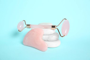 Gül kuvars yüz silindiri, gua sha aracı ve açık mavi arka planda spa taşları
