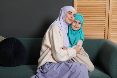 Müslüman kadın ve kızı evdeki kanepede oturuyorlar.