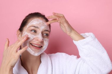 Spa günü. Güzel bir kadın pembe arka planda yüzüne maske takıyor.