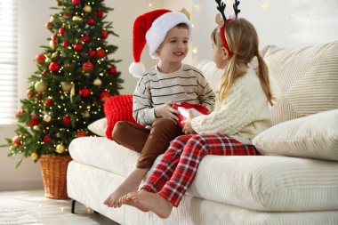 Noel Baba şapkalı küçük mutlu çocuk kız kardeşine Noel hediyesini evdeki kanepede veriyor.