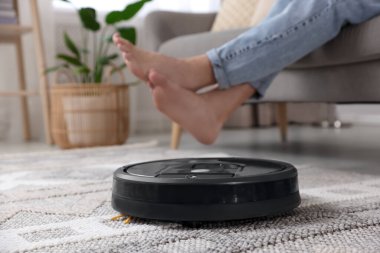 Genç adam kanepede dinlenirken robotik elektrikli süpürge evdeki yerleri süpürüyor.