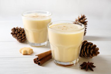 Lezzetli Noel kokteyli (eggnog) bardaklarda, kozalaklarda ve beyaz ahşap masadaki baharatlarda, yakın plan