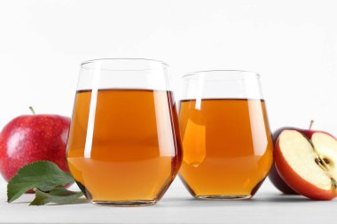 Bardaklarda taze elma suyu, meyveler ve yeşil yapraklar beyaz masada, yakın plan.