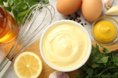 Lezzetli mayonez sosu kasede, çırpı ve malzemeler gri masada, düz yerde.