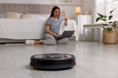 Evde dizüstü bilgisayar kullanan genç bir kadın. Robot elektrik süpürgesine odaklan.