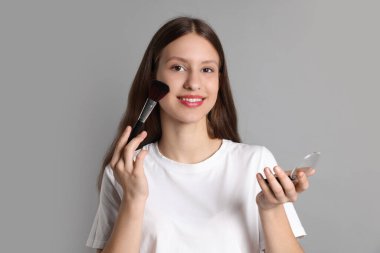 Gülümseyen genç kız gri arka planda makyaj fırçasıyla allık sürüyor.