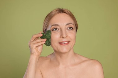 Yeşil arka planda gua sha aracıyla yüz masajı yapan güzel bir kadın.