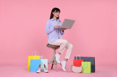 İnternet alışverişi. Dizüstü bilgisayarlı mutlu kadın pembe arka planda renkli çantaların arasında oturuyor.