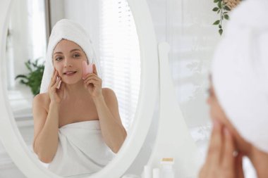Gülümseyen kadın tuvaletteki aynanın yanında gua sha aletiyle yüz masajı yapıyor.