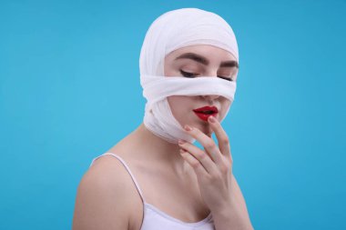 Açık mavi arka planda estetik ameliyat sonrası burnu tıbbi bandaja sarılı bir kadın.